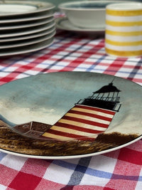 A close-up of a ceramic plate featuring a lighthouse illustration, resting on a red, white, and blue checkered tablecloth with other dishes blurred in the background.
