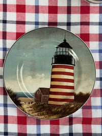 A decorative ceramic plate featuring an illustration of a red and white striped lighthouse next to a small house under a cloudy sky, displayed on a red, white, and blue checkered tablecloth.