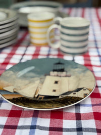 A decorative ceramic plate featuring a lighthouse and sailboat scene, displayed on a red, white, and blue checkered tablecloth with other striped dishes in the background.