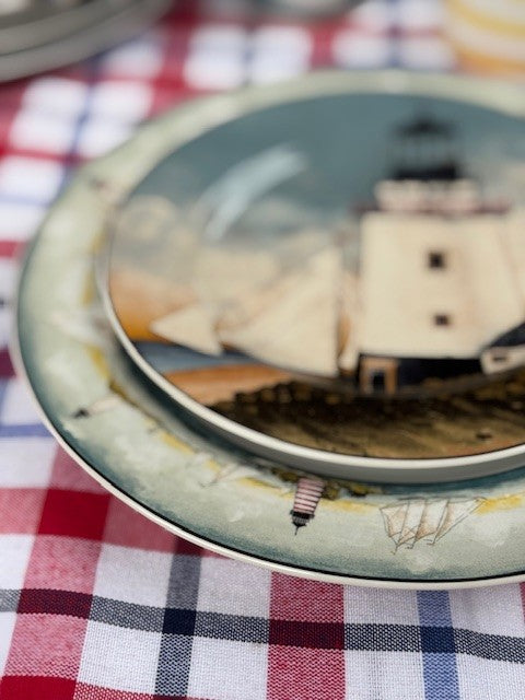 A close-up of a decorative ceramic plate featuring a lighthouse and sailboat scene, stacked on another plate over a red, white, and navy checkered tablecloth.