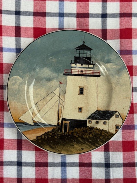 A round decorative plate featuring a painted lighthouse scene with a sailboat and small house, placed on a red, white, and blue plaid fabric background.