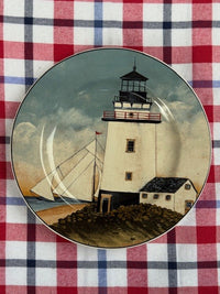 A round decorative plate featuring a painted lighthouse scene with a sailboat and small house, placed on a red, white, and blue plaid fabric background.