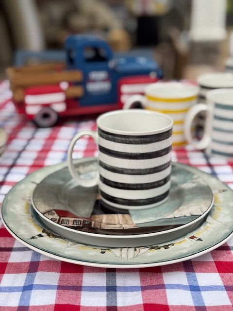 A striped ceramic mug sits on a matching saucer and plate set, placed on a red, white, and blue checkered tablecloth. A small blue and red toy truck is blurred in the background.