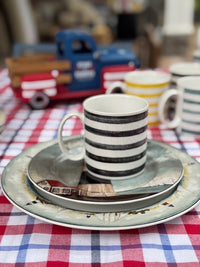 A striped ceramic mug sits on a matching saucer and plate set, placed on a red, white, and blue checkered tablecloth. A small blue and red toy truck is blurred in the background.
