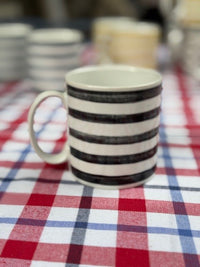 A ceramic mug with black and white horizontal stripes sits on a red, white, and blue plaid tablecloth.