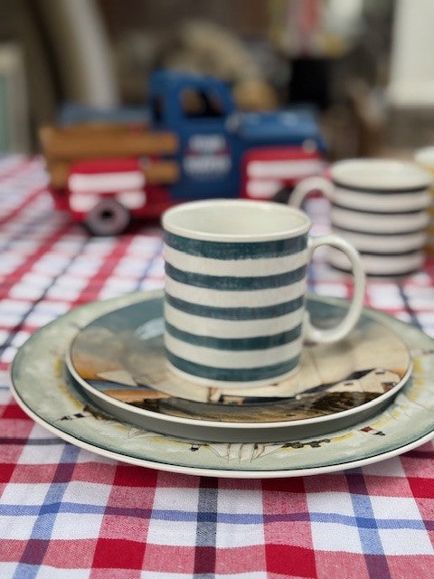 A striped ceramic mug with a handle sits on a patterned plate and saucer, placed on a red, white, and blue checkered tablecloth. A blurred toy truck and another stacked mug are visible in the background.