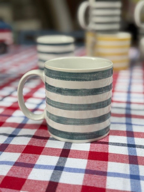 A white ceramic mug with horizontal dark teal-blue stripes sits on a red, white, and blue checkered tablecloth. Other striped mugs are blurred in the background.