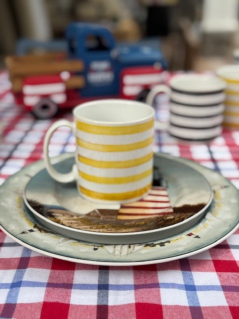 A white ceramic mug with yellow horizontal stripes sits on a decorative plate and saucer set on a red, white, and blue checkered tablecloth. A blurred blue toy truck and stacked striped bowls are visible in the background.