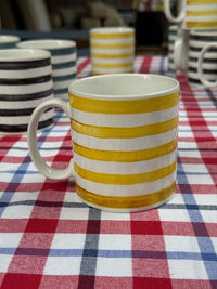 A white ceramic mug with bold yellow horizontal stripes sits on a red, white, and blue checkered tablecloth, with other striped mugs blurred in the background.