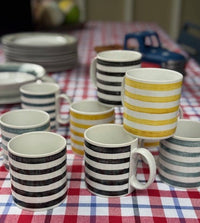 A group of ceramic mugs with horizontal stripes in black, yellow, and blue, arranged on a red, white, and blue checkered tablecloth.