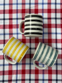 Three ceramic mugs with striped patterns are arranged on a red, white, and blue plaid fabric background. One mug has black and white stripes, while the other two have yellow and white stripes and teal and white stripes.