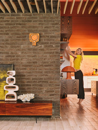 brick exposed wall with stoneware wall sculpture, woman in yellow t-shirt reaching for vase on shelf and coral colored wicker mushroom lamp