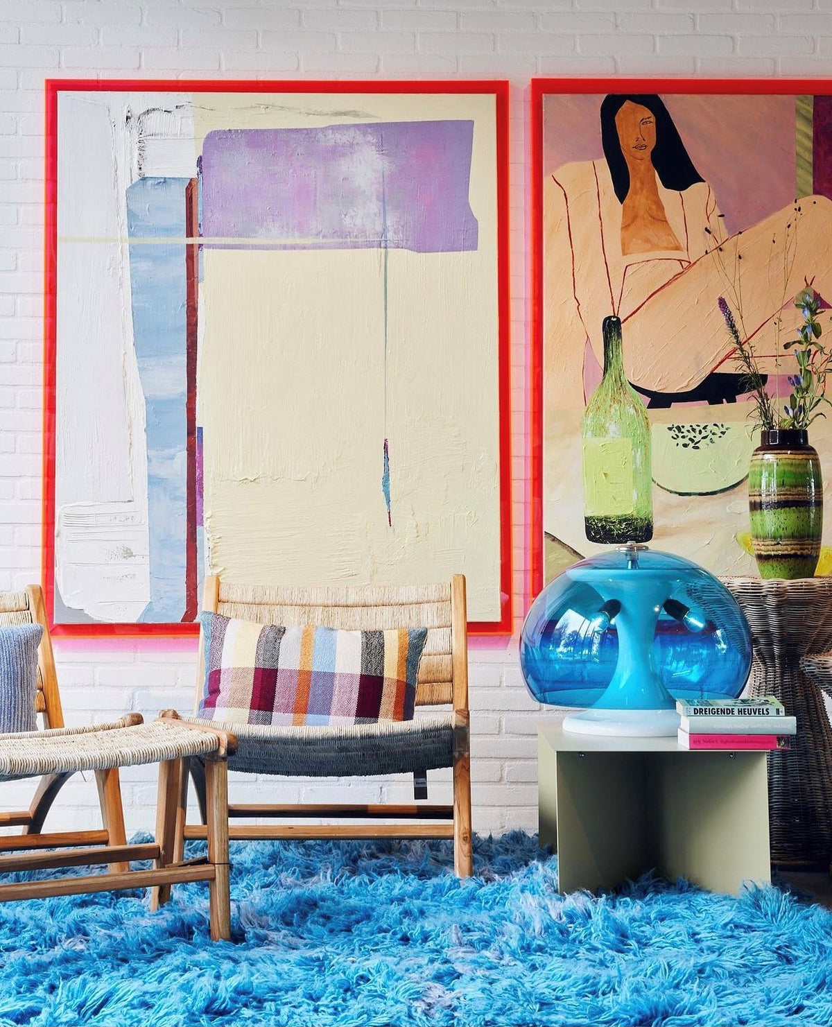 two large abstract paintings in pink acrylic frames on a wall with a blue area rug, rattan chairs and a blue lamp