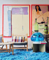 two large abstract paintings in pink acrylic frames on a wall with a blue area rug, rattan chairs and a blue lamp
