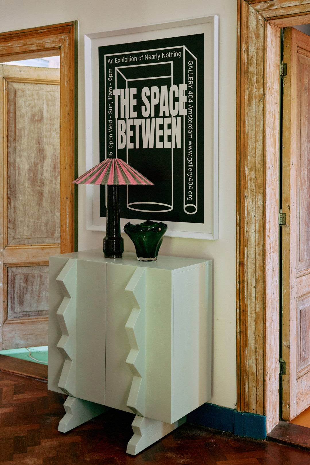 Decorative cabinet with a lamp and vase against a wall with a framed poster.