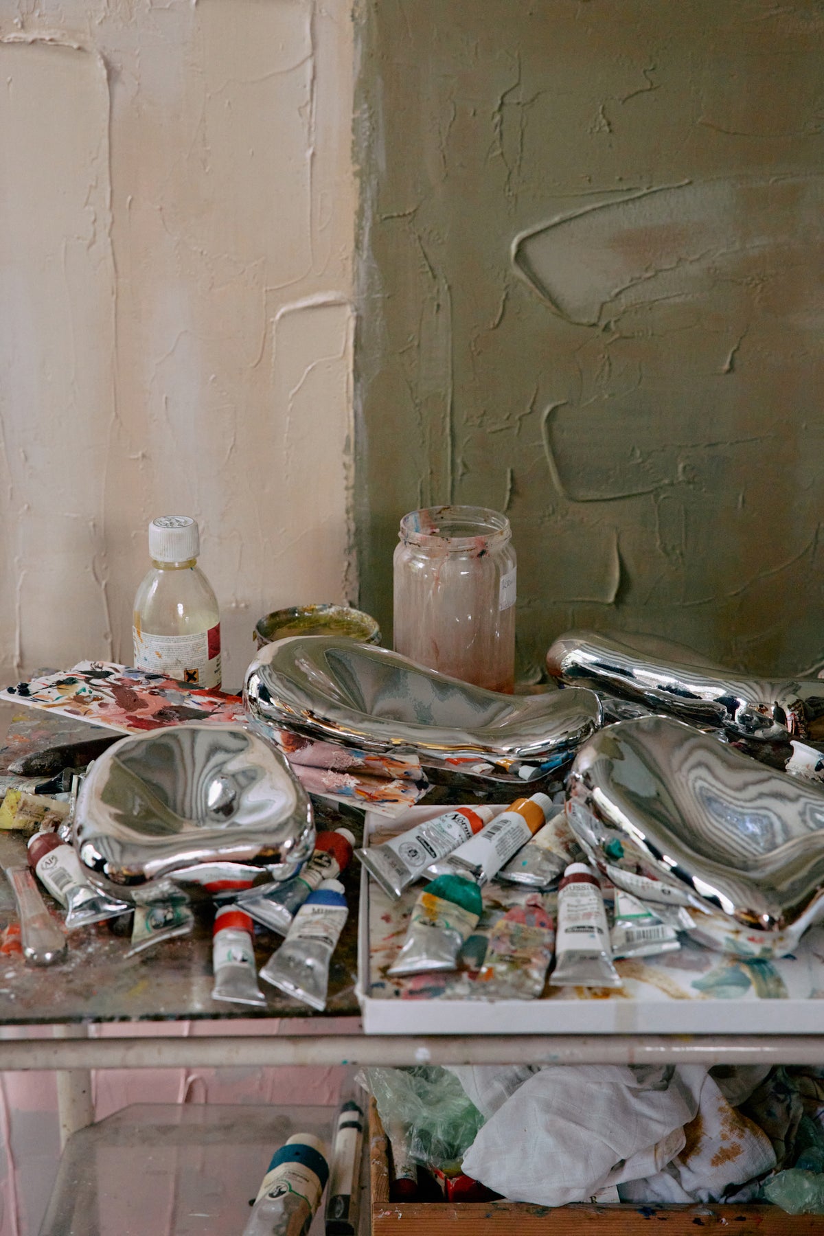 Art supplies including paint tubes, brushes, and jars on a table with a textured wall background.