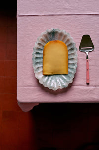 Rectangular piece of cheese on a decorative plate with a pink spatula on a pink surface.