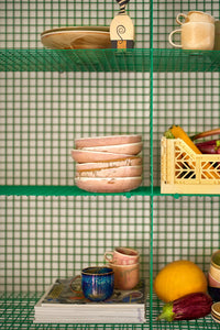 Green metal shelving unit with ceramic bowls, mugs, and a yellow crate against a green grid background.