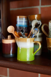 Green mug with kitchen utensils on a wooden shelf against a tiled wall.