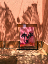Framed artwork with palm tree silhouette on a pink background, casting shadows on a textured wall.