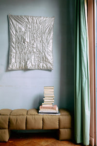 Decorative tapestry on a wall above a stack of books on a sofa with a green curtain.