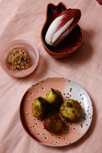 Figs on a decorative plate with a small bowl of nuts on a pink surface