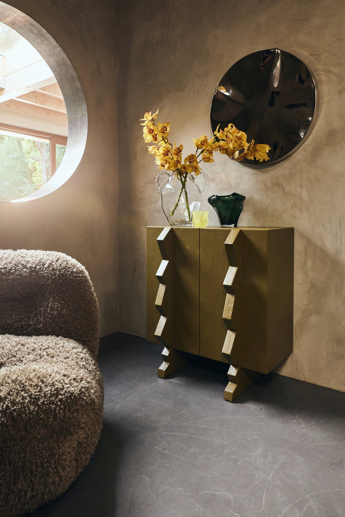 olive green wooden cabinet with sculptural handles in a moody room with glass vases and round mirror