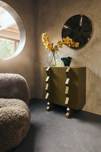 olive green wooden cabinet with sculptural handles in a moody room with glass vases and round mirror