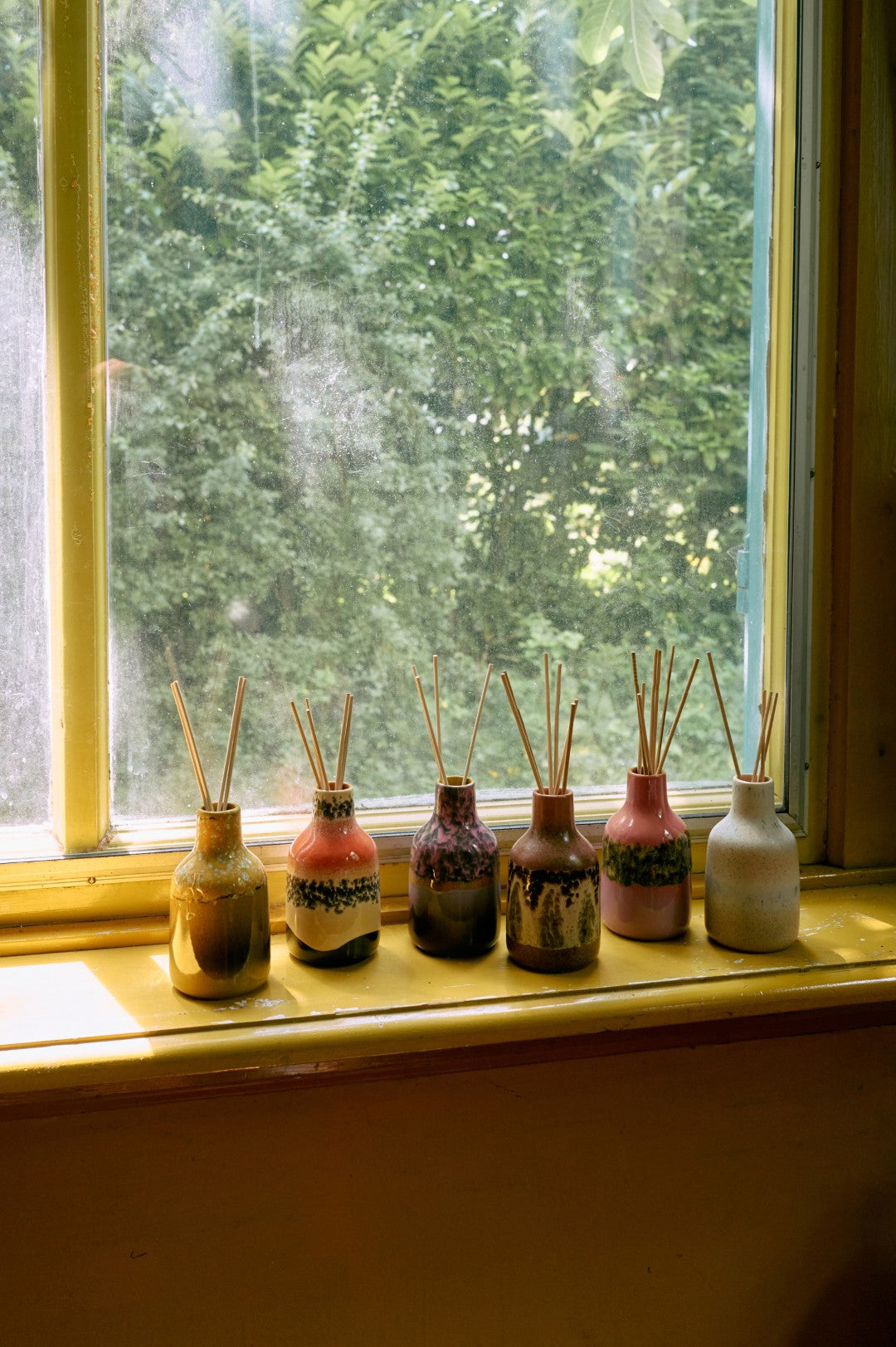 Set of diffuser bottles with sticks on a windowsill with a view of trees outside.