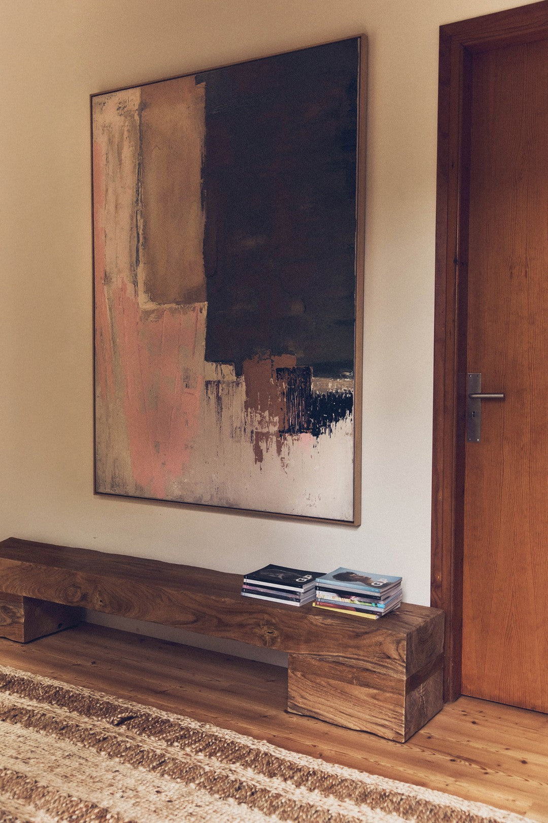 Abstract painting on a wall above a wooden bench with books, in a room with a wooden door and striped rug.