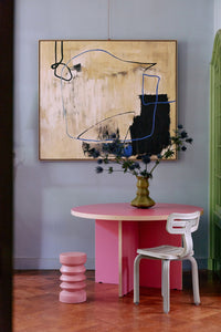 Pink table with a white chair, abstract painting on the wall, and pink cylindrical objects on a wooden floor.