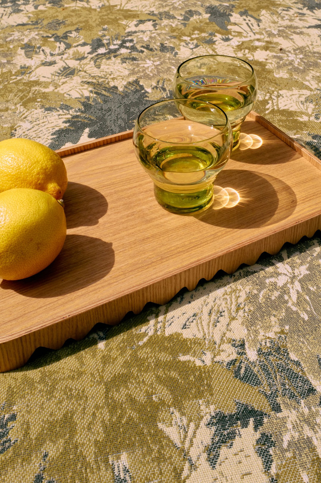 Two glasses of green liquid on a wooden tray with two lemons, all on a patterned surface.