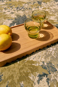 Two glasses of green liquid on a wooden tray with two lemons, all on a patterned surface.