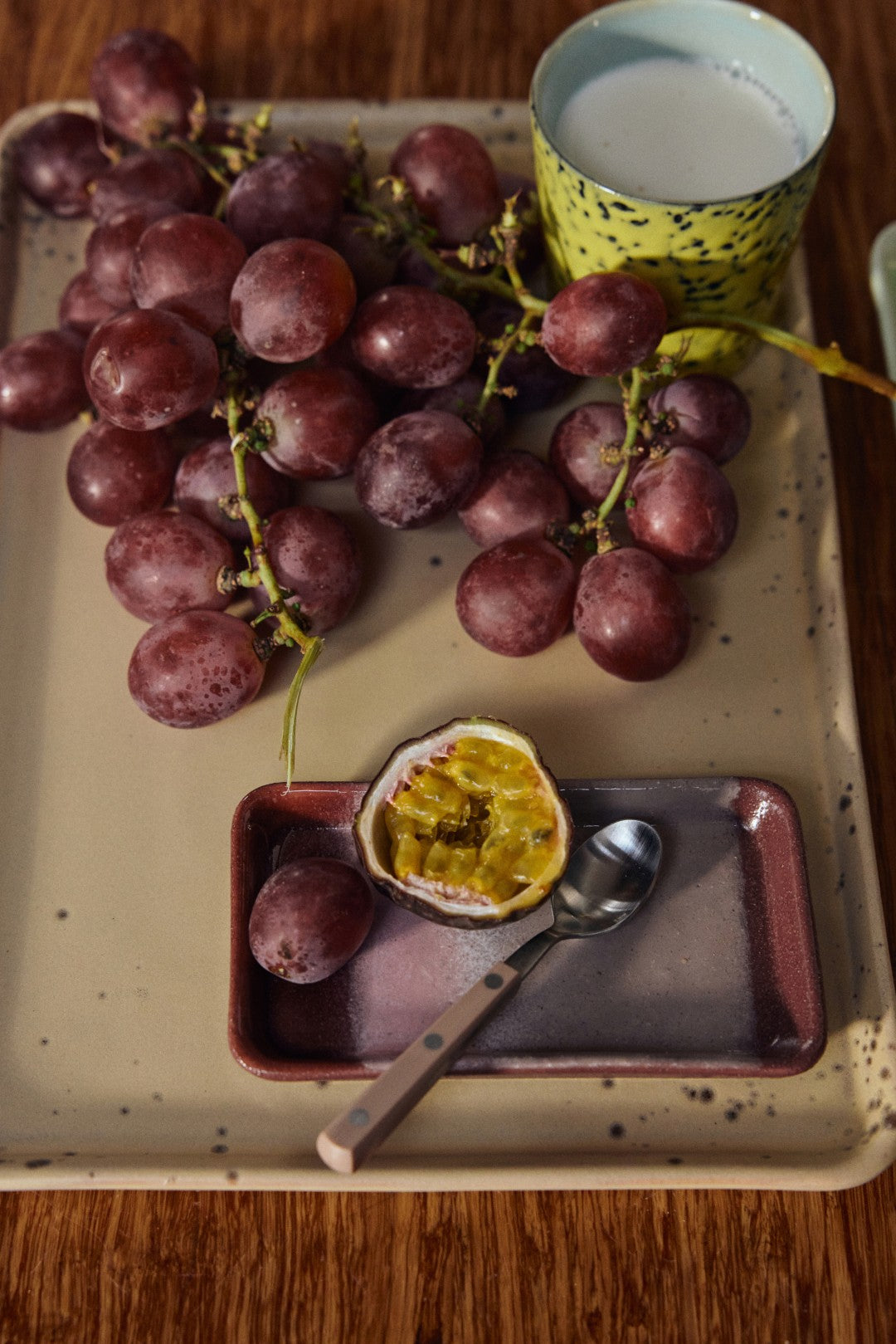 Tray with grapes, a half-eaten passion fruit, a spoon, and a cup of milk on a wooden surface.