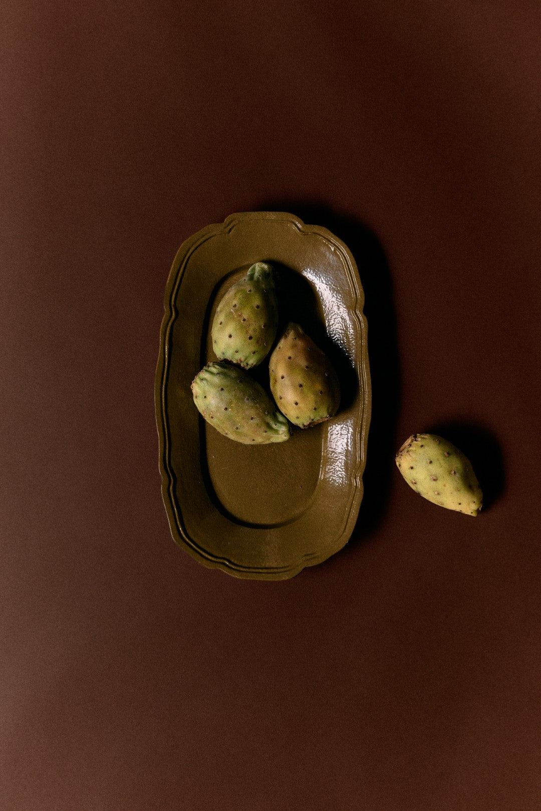 Three prickly pears on a brown ceramic plate with one outside the plate on a brown background