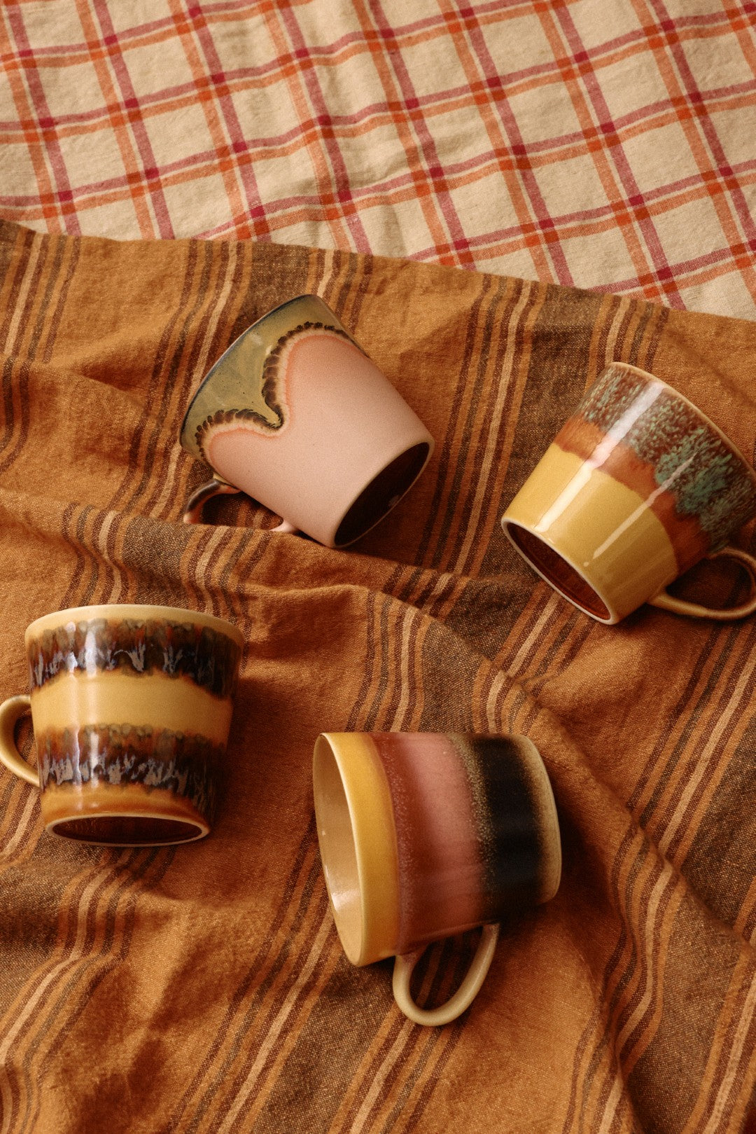 Four ceramic mugs with brown and beige glaze on a plaid fabric background