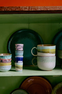 Set of colorful ceramic mugs and plates on a green shelf against a green wall.