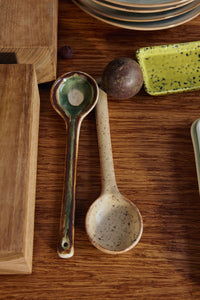 Two ceramic spoons on a wooden surface with a cutting board and fruit in the background.