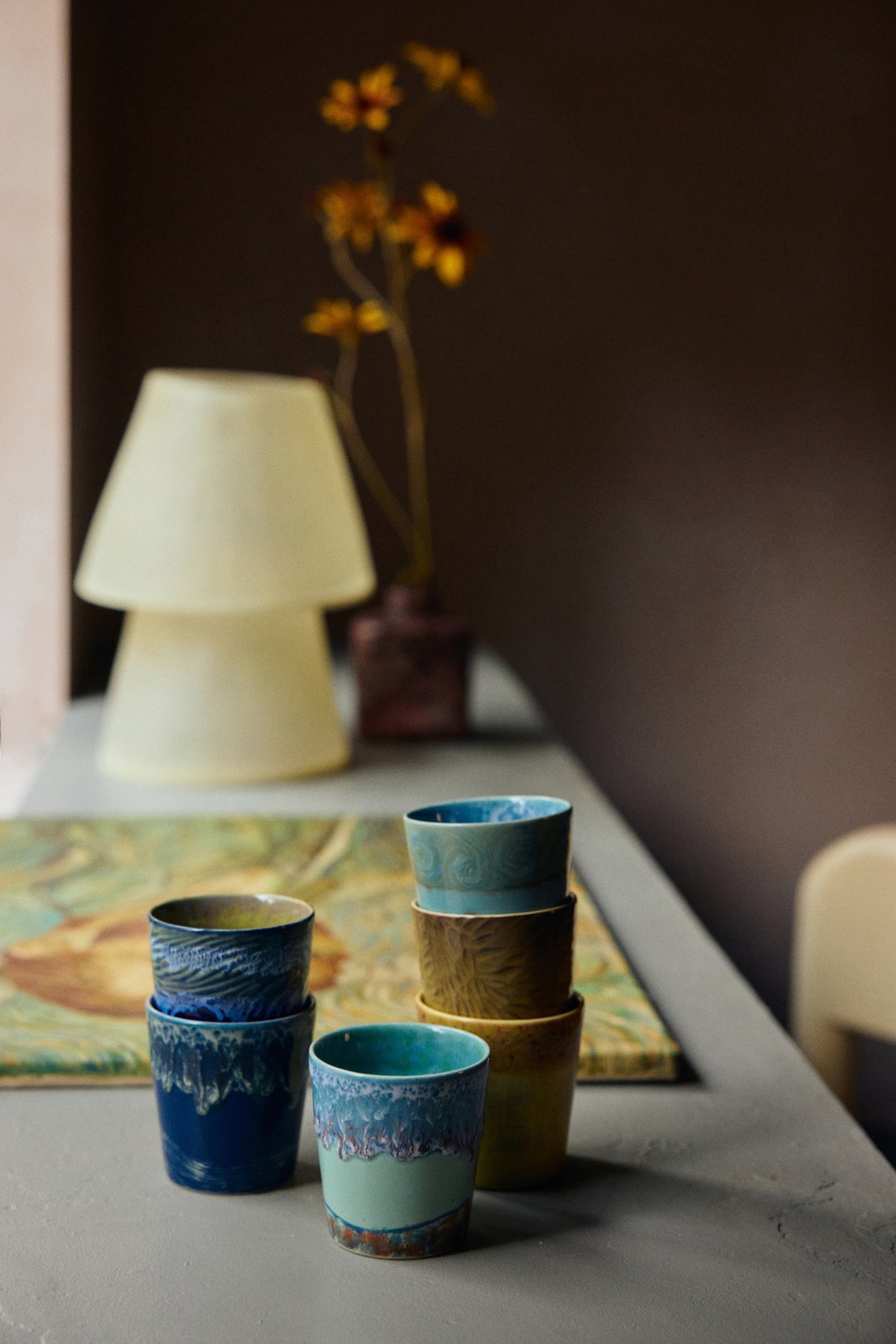 Stack of ceramic cups in blue and brown on a table with a lamp and painting in the background.