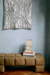Stack of books on a brown bench with a textured wall hanging above