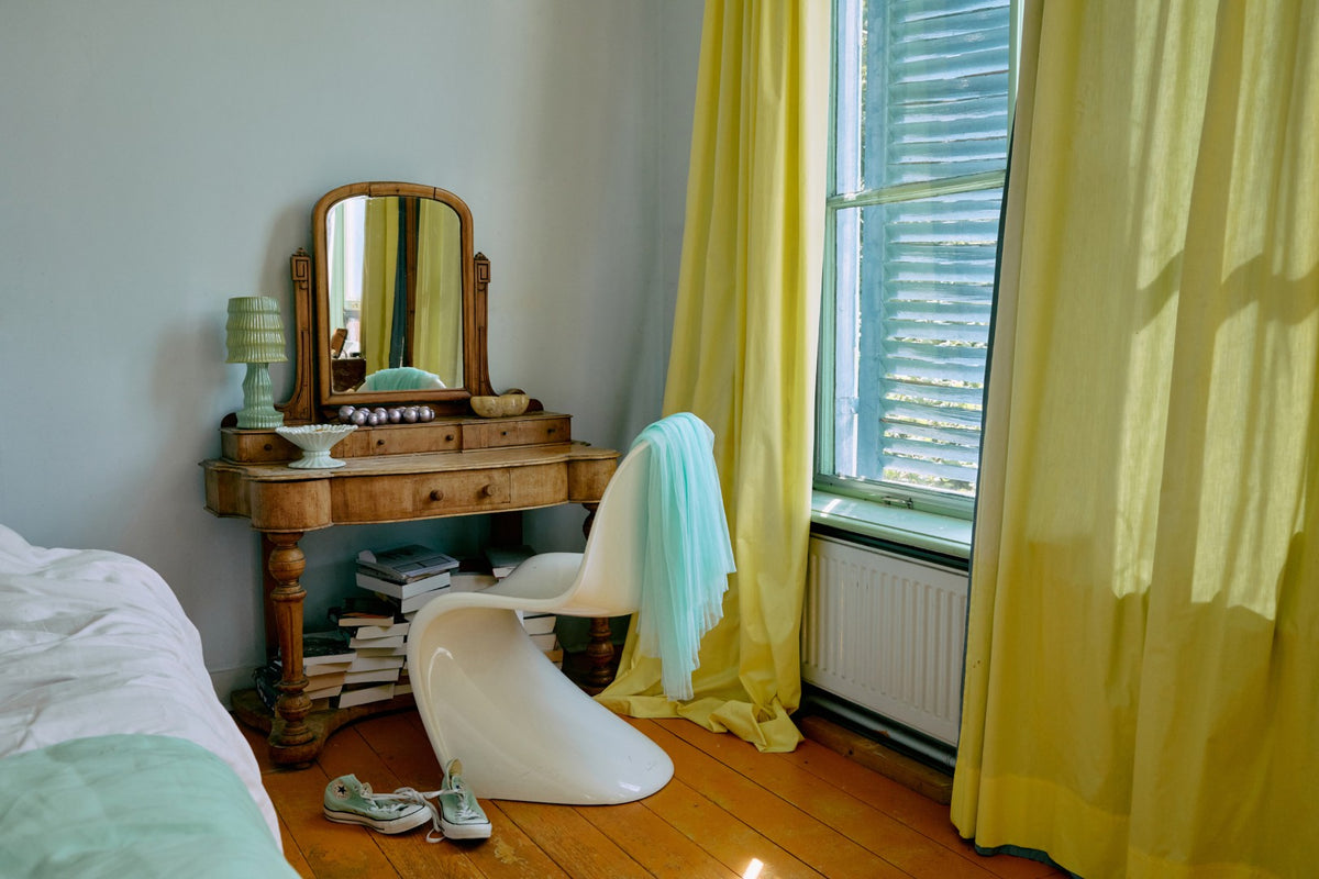 Room interior with a vanity table, mirror, and white chair near a window with yellow curtains.