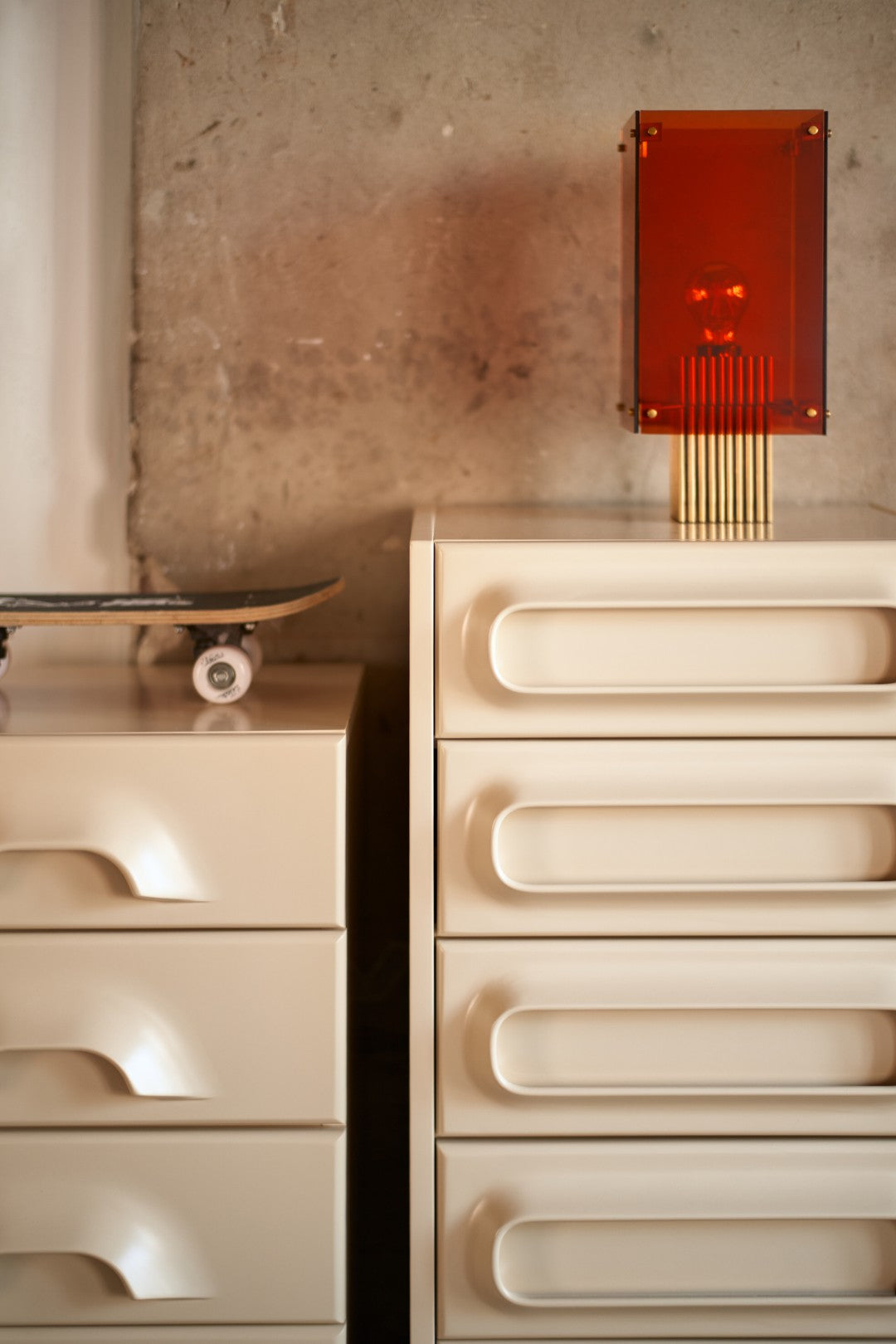 A modernist cream-colored drawer chest with curved handles stands against a textured concrete wall, topped with a translucent amber-orange rectangular table lamp with gold vertical rods. A skateboard rests on another matching cream drawer unit to the left.
