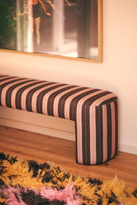 Striped bench with pink and brown pattern on a wooden floor, with a blurred painting in the background.