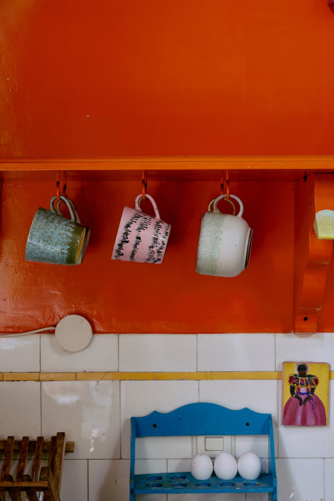 a bright orange wall with a wooden shelf and 3 mugs hanging from it in a kitchen 