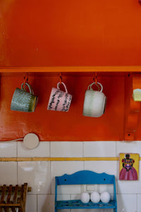 a bright orange wall with a wooden shelf and 3 mugs hanging from it in a kitchen 