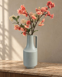 A light green ceramic vase with a handle, filled with pink and peach artificial cherry blossom branches, set on a stone countertop against a textured beige wall with soft window shadows.
