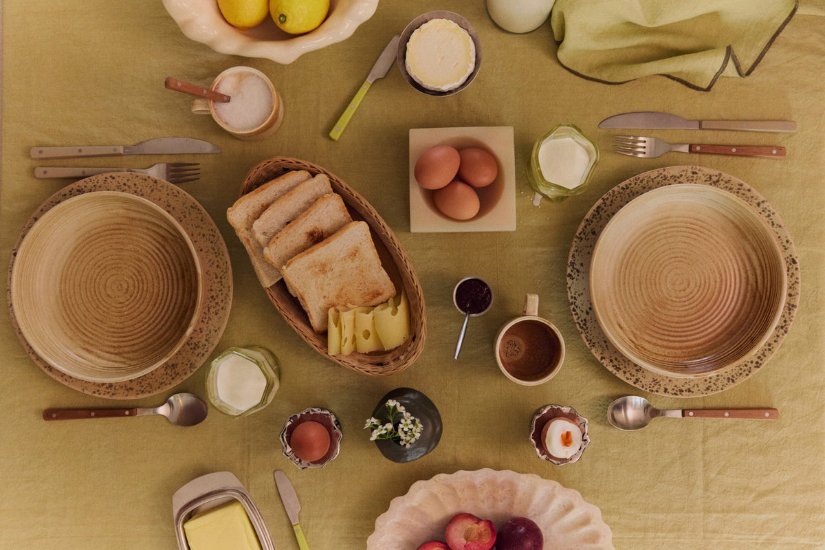 Breakfast table setting with woven plates, bread, eggs, and fruit on a yellow tablecloth.