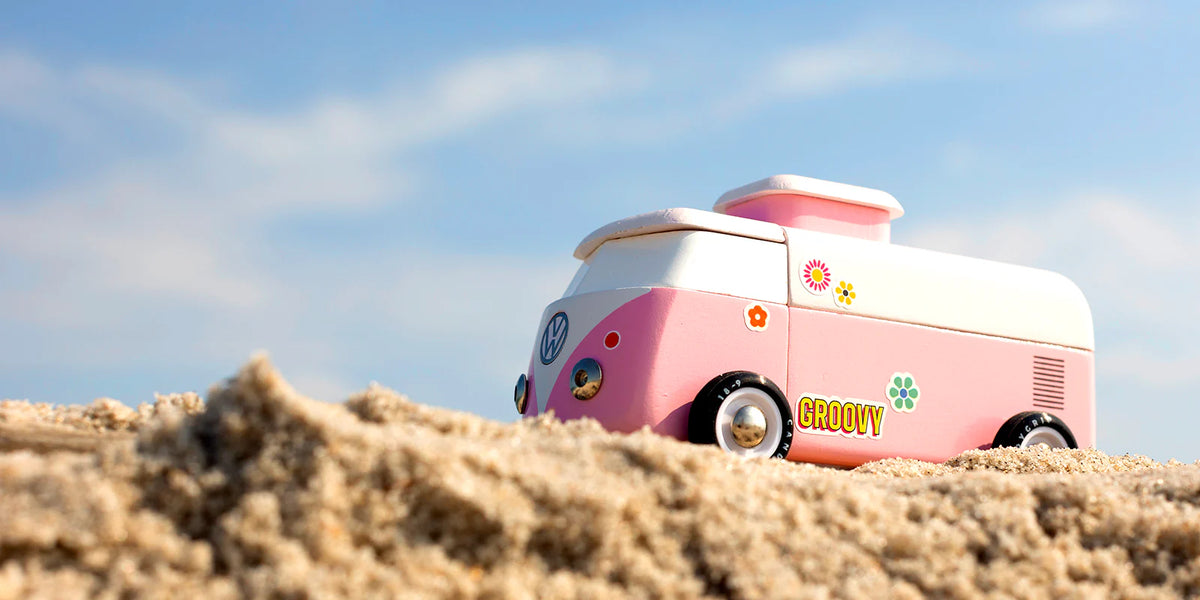 Pink and white vintage-style camper van on sandy terrain with a blue sky.