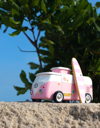 Pink and white toy van with a surfboard on sand and green foliage in the background