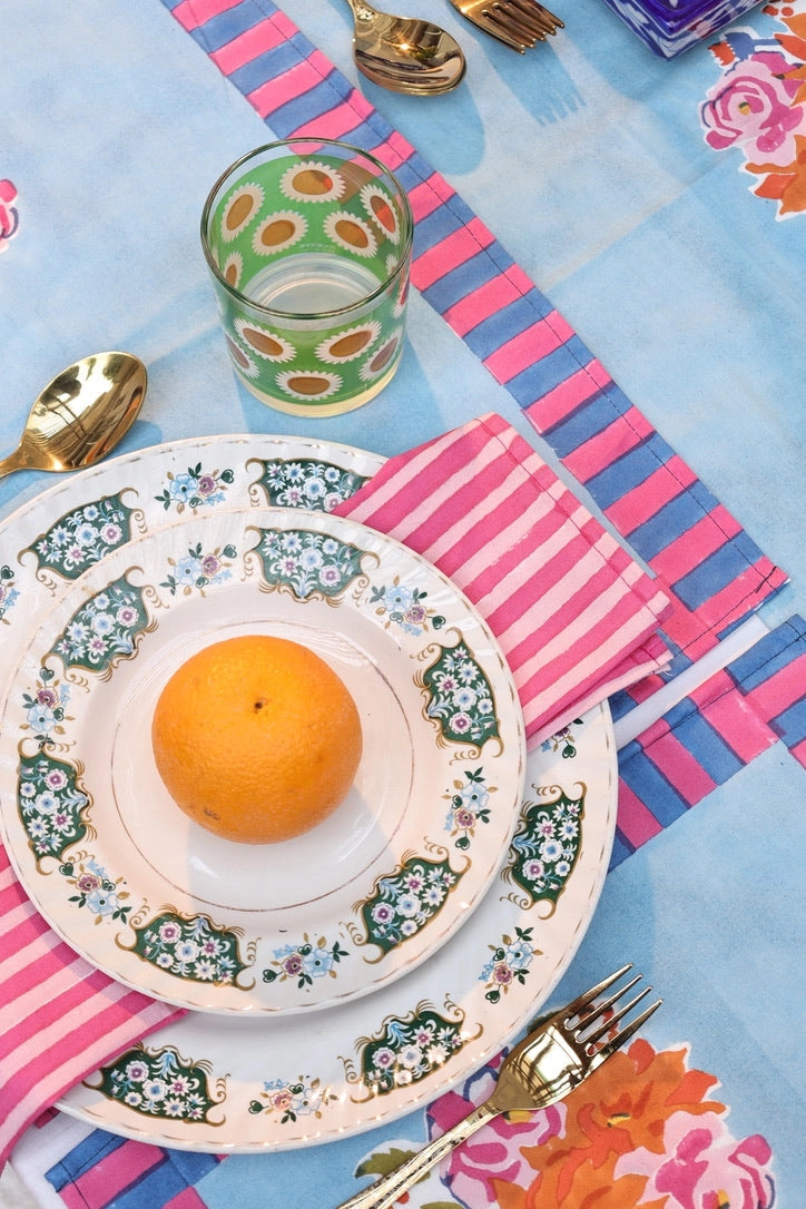 Decorative table setting with floral plates, orange, glass, and cutlery on a colorful tablecloth.